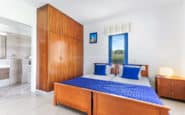 Modern minimalist bedroom with blue accents, wooden bed, wardrobe, and artwork in Daphne-16.