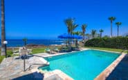 Luxurious coastal pool area with lounge chairs, umbrellas, palm trees, and serene ocean views.