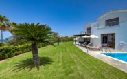 Luxurious contemporary villa for rent with palm tree, pool, and lush lawn on sunny day.