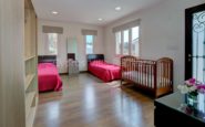 Cozy and modern bedroom with twin beds, crib, storage shelf, dresser, and natural light.