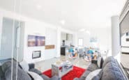 Modern living space with grey sofas, TV, fireplace, dining area, and natural light.