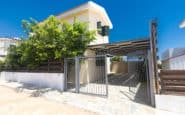 Modern suburban villa with carport, gate, and lush tree in sunny setting.